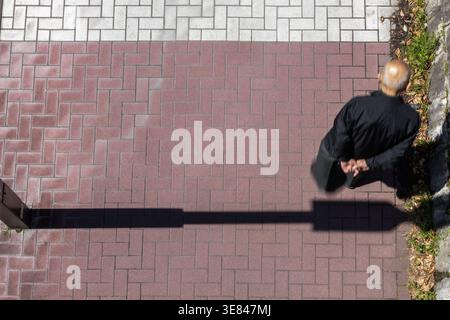 Overhead of contemplative senior man walking with hands behind back and slightly hunched forward on sunlit red brick walkway with elongated shadows. Stock Photo