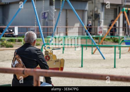 Reflective moment–solitary older man, with short grey hair sits alone in an empty playground, quietly gazing at the surroundings as people bass by. Stock Photo