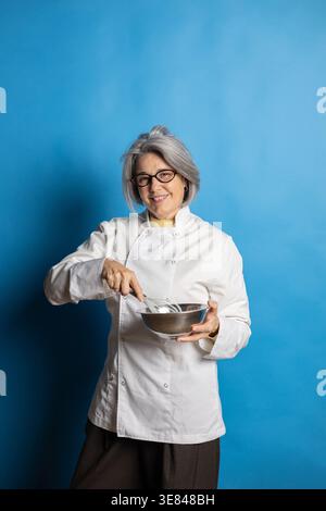 Senior woman chef smiling and whisking ingredients in a metal bowl Stock Photo