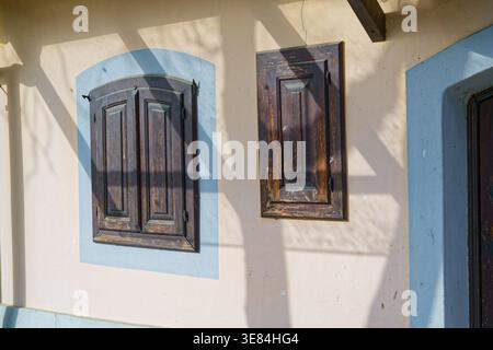 The image shows two closed wooden shutters on a light-colored wall. The shutters are dark brown and appear weathered. Sunlight casts shadows on the wa Stock Photo