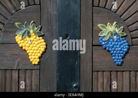 Decorative ceramic grapes, one yellow and one blue, adorn a rustic wooden door. These charming embellishments likely serve as a welcoming touch, hinti Stock Photo