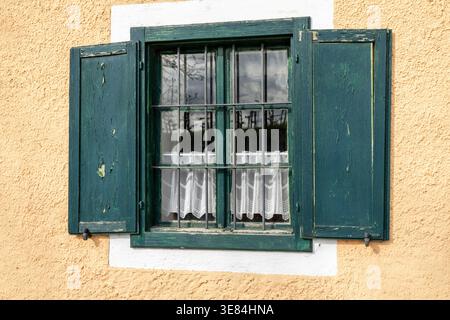 A weathered, teal-painted wooden shutter is open, revealing a window with iron bars and delicate white curtains. The window is set into a textured, pa Stock Photo