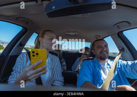 Family on summer road trip in a car: father driving, mother in passenger seat using smartphone, two kids in back entertained by devices, all smiling a Stock Photo