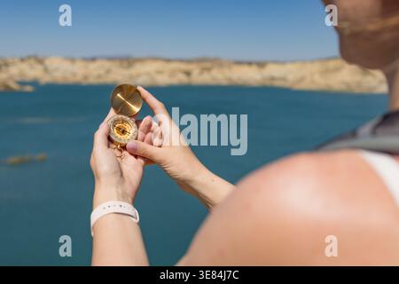 Woman standing outdoors by a vast lake and rocky terrain, holding a vintage golden compass in her hands, carefully checking the directions for navigat Stock Photo