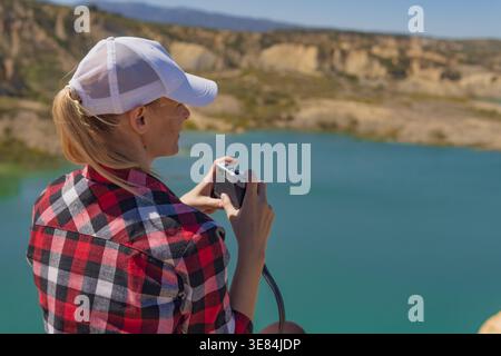 Young woman wearing a white cap and plaid flannel shirt capturing scenic views of a vibrant turquoise lake and rugged badlands with a camera while exp Stock Photo
