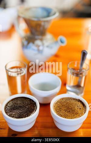 Two white cups of coffee with crema on a wooden table, flanked by two water glasses and an empty bowl, with a blurred pour-over dripper and kettle in Stock Photo