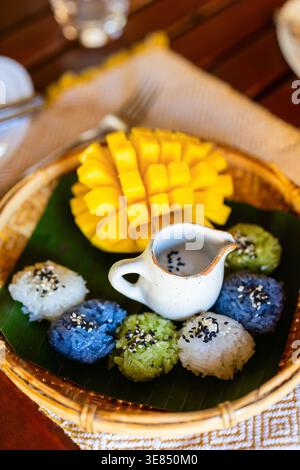 Colorful Thai-style sticky rice dessert with mango and coconut milk sauce served on banana leaf in woven tray, top-down close-up on wooden table. Stock Photo