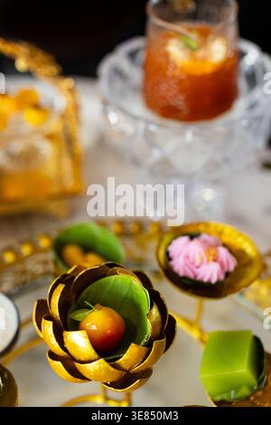 Luxury afternoon tea set on marble table with ornate gold stand holding assorted bite-size desserts and fruit, with iced tea drinks and bokeh cafe bac Stock Photo