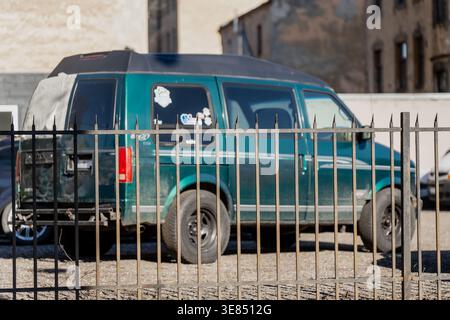 Old green cargo van behind iron fence on gravel yard with weathered urban buildings, delivery transport, repair fleet and local business work theme Stock Photo