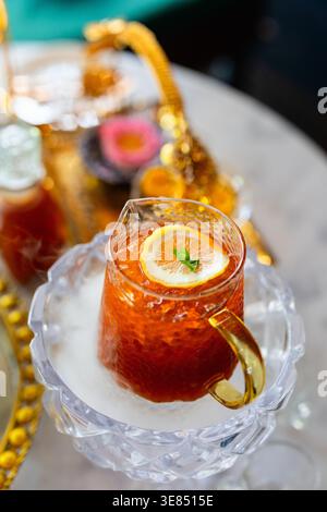 Glass cup of iced tea with lemon slice and mint on crystal saucer, gold handle, on cafe table with ornate teapot tray background bokeh. Stock Photo