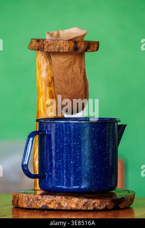 Rustic cloth coffee filter dripper on wooden stand over blue speckled enamel mug on table, green background. Handmade pour-over brewing, cozy vintage Stock Photo