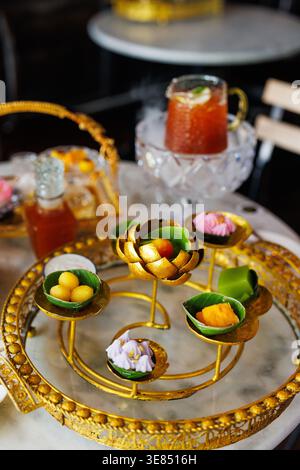 Luxury afternoon tea set on marble table with ornate gold stand holding assorted bite-size desserts and fruit, with iced tea drinks and bokeh cafe bac Stock Photo