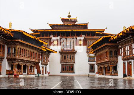 Symmetrical view of traditional bhutanese dzong monastery courtyard with ornate wooden balconies and golden roofs, wet stone paving after rain, overca Stock Photo