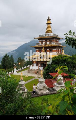 Ornate buddhist temple with golden roof and spire, centered in lush hillside garden, misty mountain forest backdrop under cloudy sky. Stock Photo