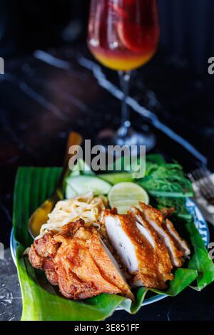Thai fried chicken served on banana leaf with cucumber, cabbage, lime, and herbs; colorful cocktail in background on dark marble table, shallow depth Stock Photo