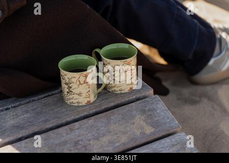 Two ceramic mugs with hot drink on weathered wooden bench beside seated person on beach sand, cozy outdoor break and seaside relaxation concept Stock Photo