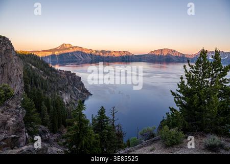 View of a serene blue lake reflecting the soft glow of the sunrise, surrounded by towering cliffs and lush green forests, Crater Lake, Oregon, United Stock Photo