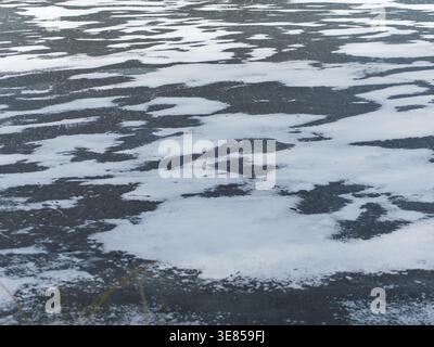 A detailed top-down view of a frozen water surface during winter. Dark, smooth ice is partially covered with irregular patterns. Stock Photo