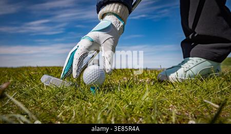 Low angle view of golfer hand placing golf ball on tee with club on grassy course during sunny day. Stock Photo