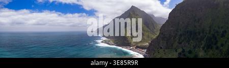 Panoramic view of Madeira Island north coast shows jagged cliffs, Atlantic surf along a narrow shore, and a small settlement at the base under driftin Stock Photo