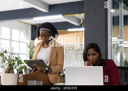 Diverse female coworkers collaborating in business wear at open office using tablet and laptop Stock Photo