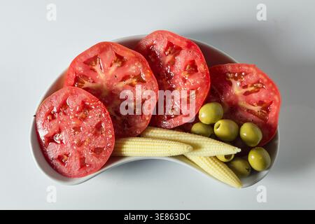 A clean, top-down studio shot of fresh tomato slices, pickled baby corn, and green olives on a white kidney-shaped plate. Bright, high-key lighting em Stock Photo