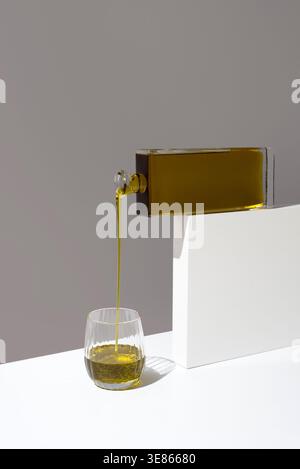 Minimalist studio still life of olive oil being poured from a rectangular glass bottle into a tumbler on white blocks, capturing tasting, flavor and g Stock Photo
