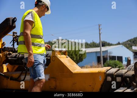 Male engineering worker in hard hat and reflective vest inspects yellow construction machine outdoors at an industrial site, focusing on maintenance, Stock Photo