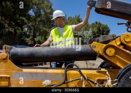 Male construction worker in hard hat and safety vest inspects an excavator hydraulic cylinder on a sunny outdoor site, highlighting engineering, maint Stock Photo