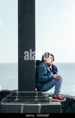 A pensive young man sits on a stone base beside a tall concrete pillar at the seaside, gazing away in quiet thought, with ocean horizon and copy space Stock Photo