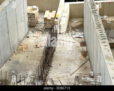 Concrete foundation under construction with exposed steel rebar cage in a trench, forming walls and floor slab. Building site shows formwork, bricks, Stock Photo