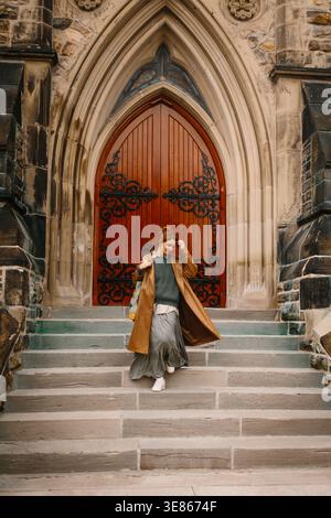 Young woman in a long coat and skirt walks down stone steps in front of an ornate gothic arched wooden church door, captured in warm natural light. Stock Photo