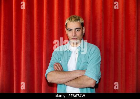 Confident young man with short dyed hair stands with arms crossed, wearing a teal shirt over a white tee, posing against a red theater curtain backdro Stock Photo