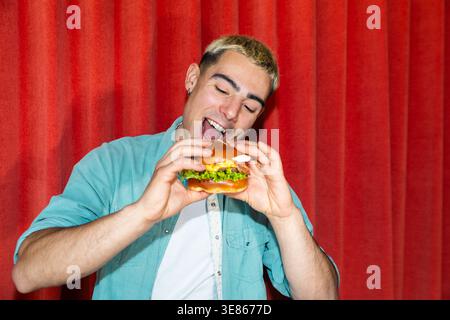 Smiling young man taking a big bite of a hamburger, holding a fresh burger with lettuce and sauce, posing playfully in front of a vivid red curtain ba Stock Photo