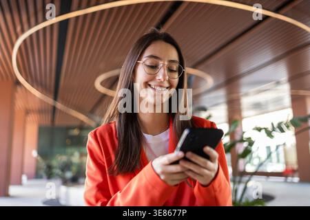 A young businesswoman in an orange blazer smiles while using her smartphone inside a modern office building, showcasing a professional and cheerful wo Stock Photo