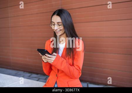 Smiling woman wearing an orange blazer and glasses uses a smartphone. She's leaning against a modern exterior wall, suggesting a dynamic business envi Stock Photo