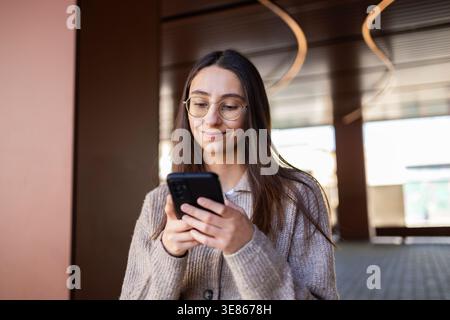 A young woman wearing glasses checks her smartphone while smiling in a modern business setting. She is standing indoors, exuding confidence and profes Stock Photo