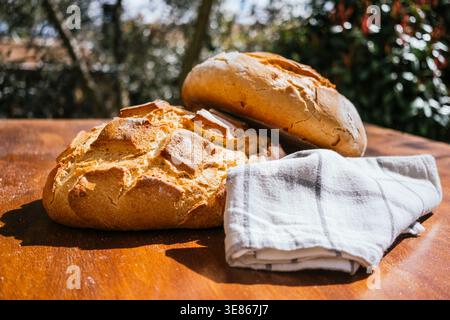 Two large, rustic village loaves rest on a wooden table outdoors, accompanied by a plaid cloth. Captured in natural light, this image evokes a sense o Stock Photo