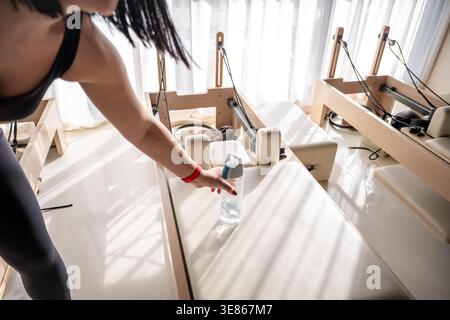 Fit woman takes a short break during pilates reformer workout, reaching for a bottle of water in a bright studio, focusing on hydration, wellness and Stock Photo