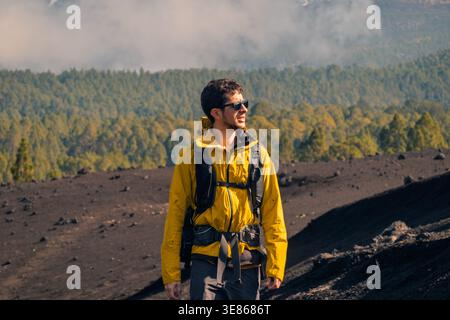 Adventurous male hiker in a yellow jacket and backpack stands on a volcanic trail, gazing over a dense forest and misty mountains under soft daylight. Stock Photo