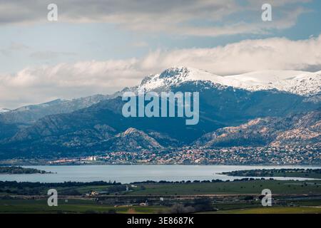 Panoramic view of snow-capped mountains rising above a calm lake, with a valley town and green farmland in the foreground under a cloudy sky. Stock Photo