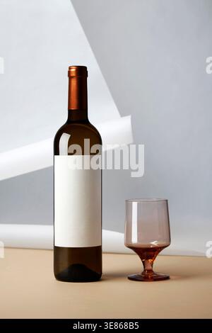 Minimal still life of a dark wine bottle with blank label and a stemless glass holding red wine, set against geometric shadows and a soft neutral back Stock Photo