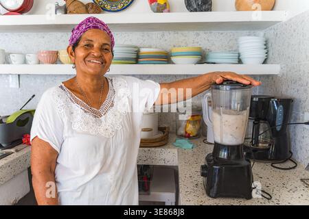 Elderly Caribbean woman using a blender in a cozy kitchen adorned with colorful dishes, creating a tropical recipe. Her vibrant headscarf adds charm t Stock Photo