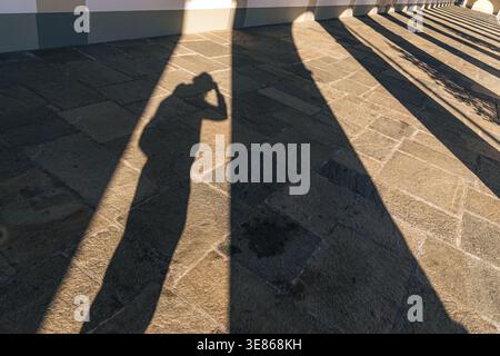 Morning sun casts long shadows of arches and a person on a tiled stone walkway in Piamonte, Italy. The serene scene captures the beauty of light and s Stock Photo