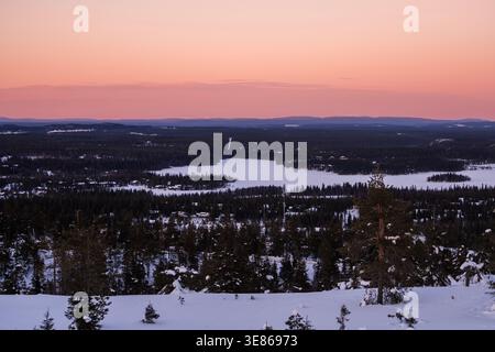 Panoramic view over snowy pine forest and frozen lake at dusk in Finland, with soft pink sky, distant hills and serene wilderness atmosphere in winter Stock Photo