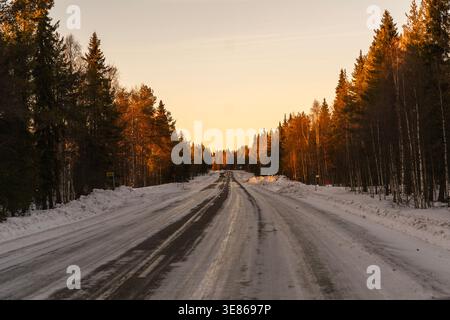 Icy winter road stretches through a snow-covered boreal forest at golden sunset, with pine and birch trees lining the quiet highway in Finland. Stock Photo