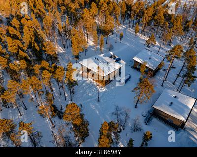 Aerial drone view of cozy wooden cabins surrounded by snow-covered pine forest in Finland, glowing in warm low winter sunlight, quiet wilderness retre Stock Photo