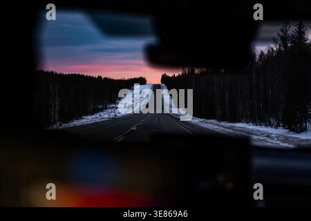 Empty winter road stretches through dark conifer forest in Finland, seen from a car, with snowy shoulders and a pink dusk sky, evoking solitude and tr Stock Photo