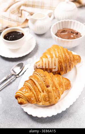 Freshly baked croissants sit on a white plate, accompanied by a cup of hot black coffee, a creamer, small sugar bowl, and a bowl of rich chocolate spr Stock Photo