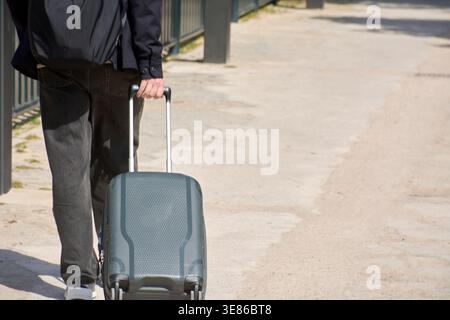 A traveler walks along a sunny paved pathway pulling a grey rolling suitcase with a telescopic handle. The person is seen from behind wearing casual d Stock Photo